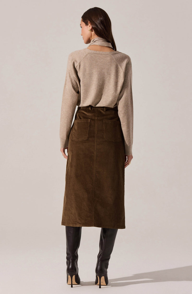 A woman is wearing a beige long-sleeve sweater with a soft, knit texture, paired with a high-waisted, dark brown corduroy midi skirt that has large patch pockets on the back. She is also wearing black, knee-high leather boots with high heels.