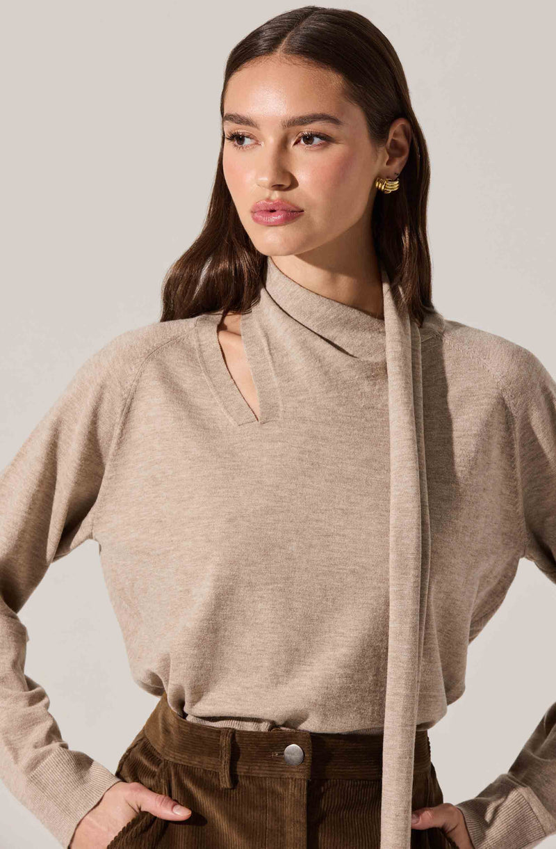 A woman is wearing a beige knit top with a distinctive neckline featuring a cutout and a long, matching scarf or tie draped over one shoulder. She pairs it with high-waisted brown pants. She accessorizes with gold hoop earrings.