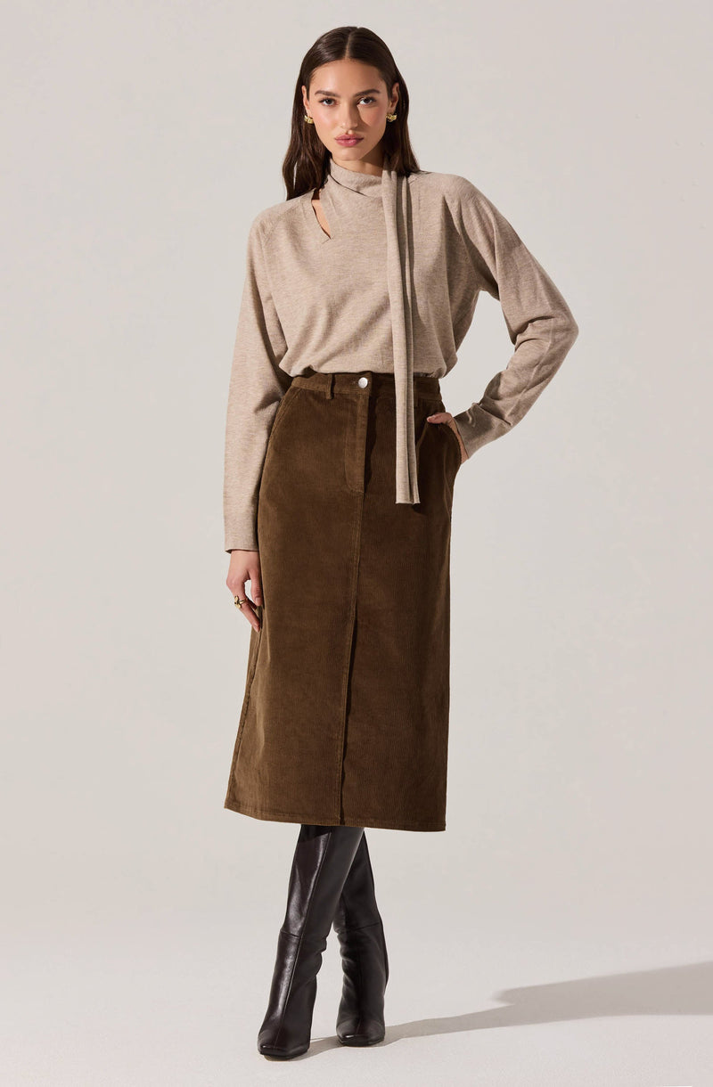 A woman is wearing a beige long-sleeved top with a high neckline and a tie detail on the side of the collar. She pairs it with a high-waisted, brown, knee-length corduroy skirt that features a front slit and a button and zip closure. She completes the outfit with black knee-high boots.