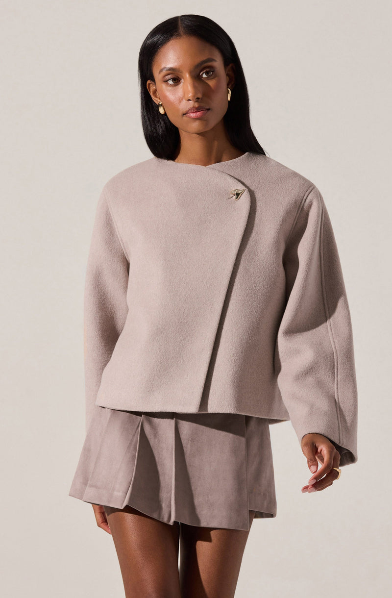 A woman is wearing a light taupe, oversized, cropped coat with a minimalist design. The coat features a single decorative pin or brooch near the neckline and has long, loose sleeves. She is also dressed in matching taupe shorts with pleats.
