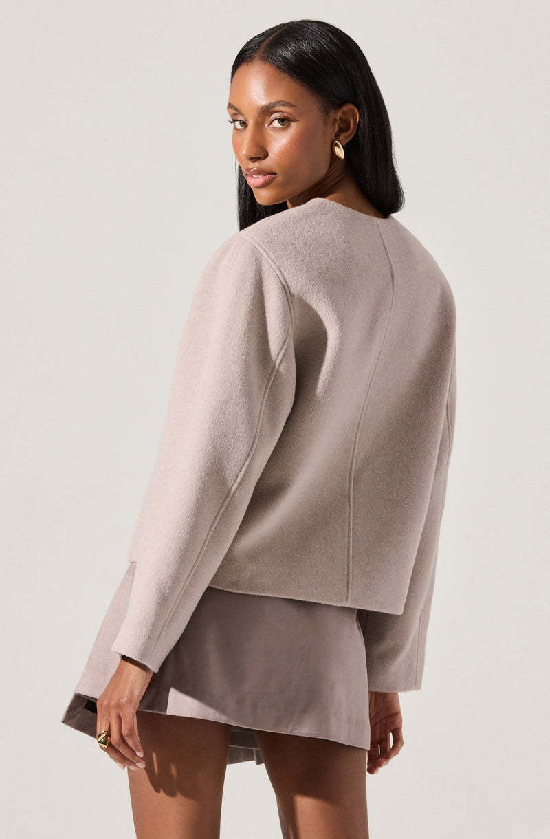 A woman is wearing a light beige, long-sleeved, slightly oversized blazer with a rounded neckline. The blazer appears to be made of a soft, textured fabric. She pairs it with a matching beige skirt or dress. The outfit has a sleek, minimalistic style.