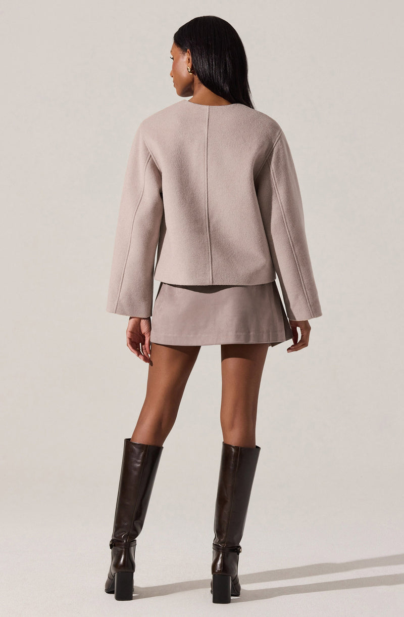 A woman is wearing a light beige, structured blazer with long, slightly flared sleeves and a subtle seam detail down the back. She pairs it with a matching mini skirt and tall, dark brown leather knee-high boots with block heels.