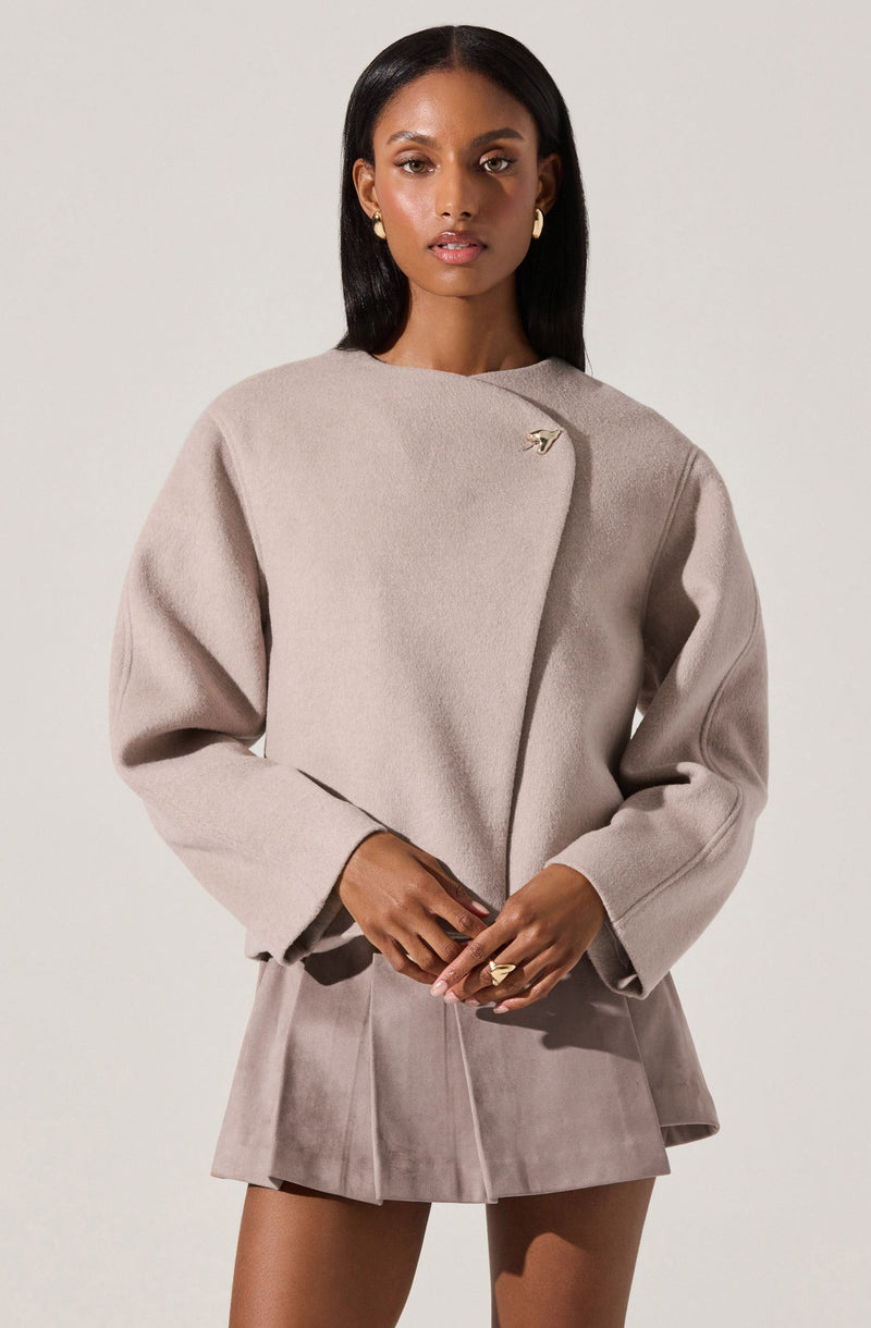 A woman is wearing a light beige, long-sleeved, structured coat with a high neckline and an asymmetrical closure secured by a decorative pin on the upper chest. She pairs it with a matching beige mini skirt with pleats, made from a soft, suede-like material. The outfit is complemented by gold earrings and rings.