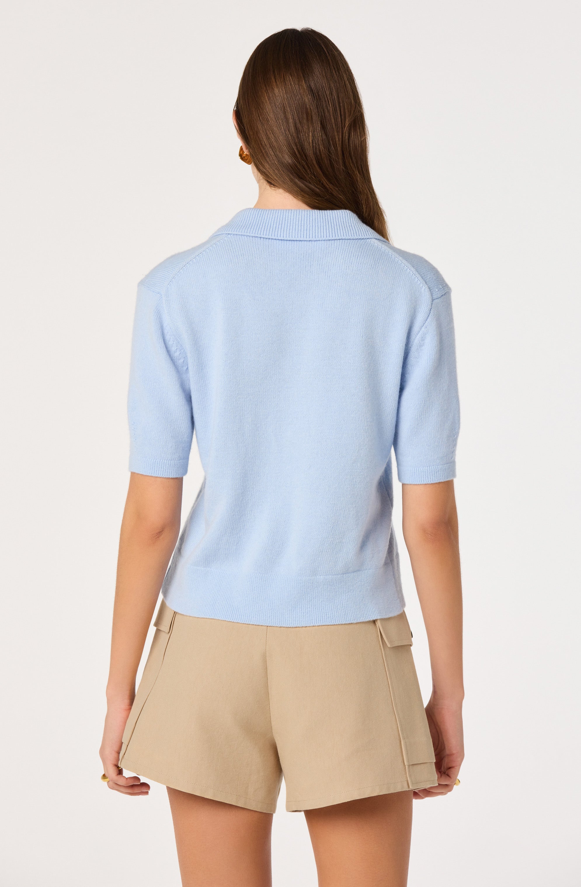Woman in a light blue short-sleeve sweater and beige shorts, back view.