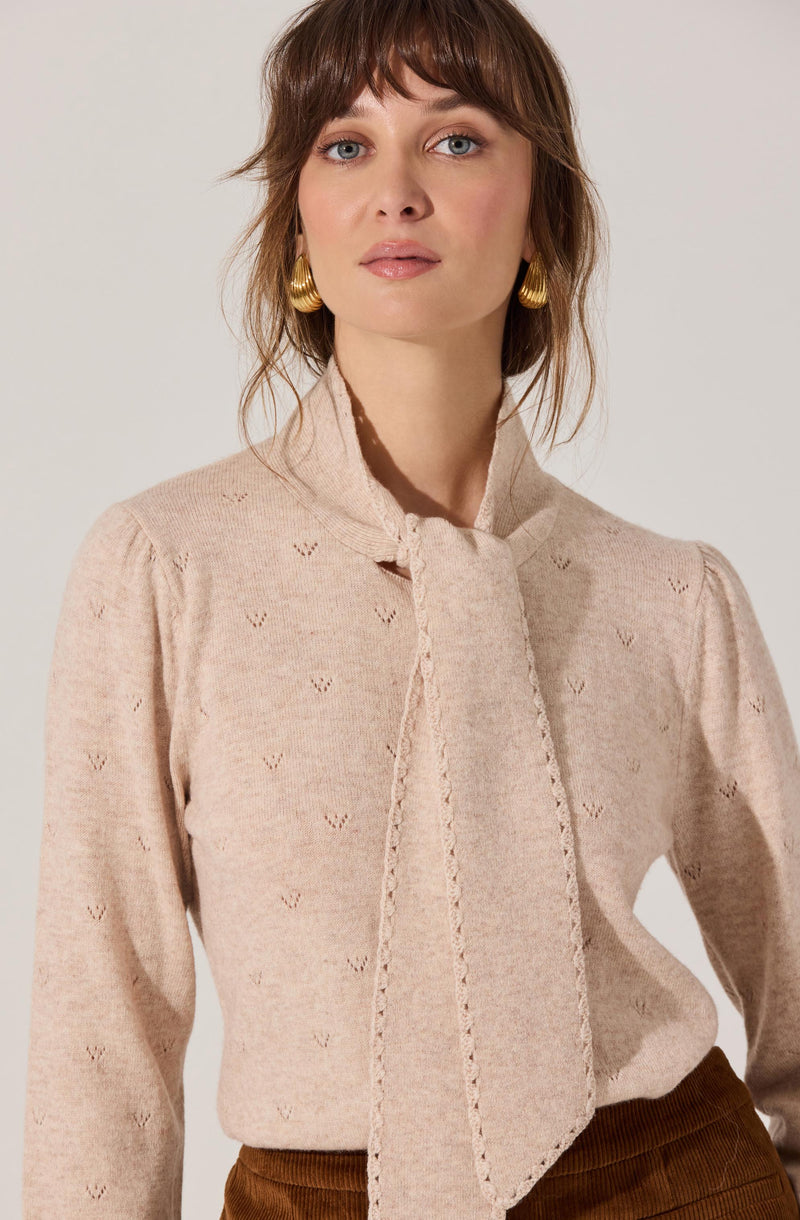 A woman is wearing a light beige sweater with a subtle embroidered pattern across the fabric, featuring small, repeated heart-like shapes. The sweater has a high neckline with a vertical tie detail at the front. She is also wearing gold hoop earrings.