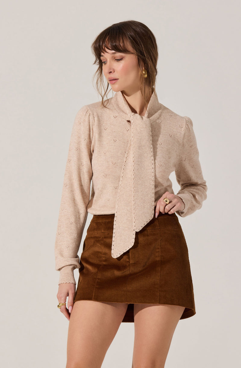 A woman is wearing a light beige long-sleeve blouse with small, subtle embroidered details. The blouse features a large, matching fabric tie at the neck that hangs down the front. She pairs it with a high-waisted, brown corduroy mini skirt.