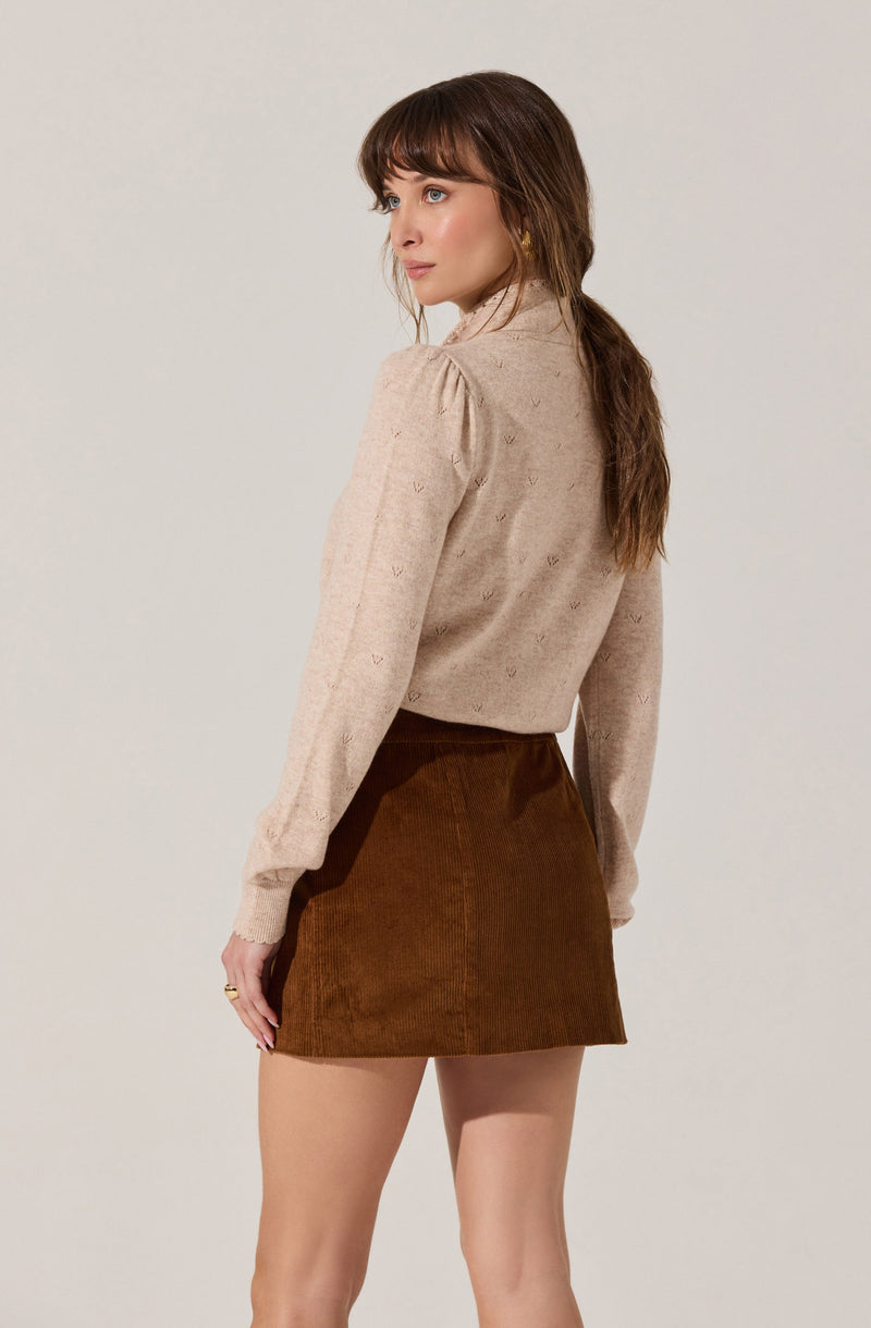 A woman is wearing a beige sweater with subtle embroidered details and puffed long sleeves, paired with a high-waisted, brown corduroy A-line mini skirt.