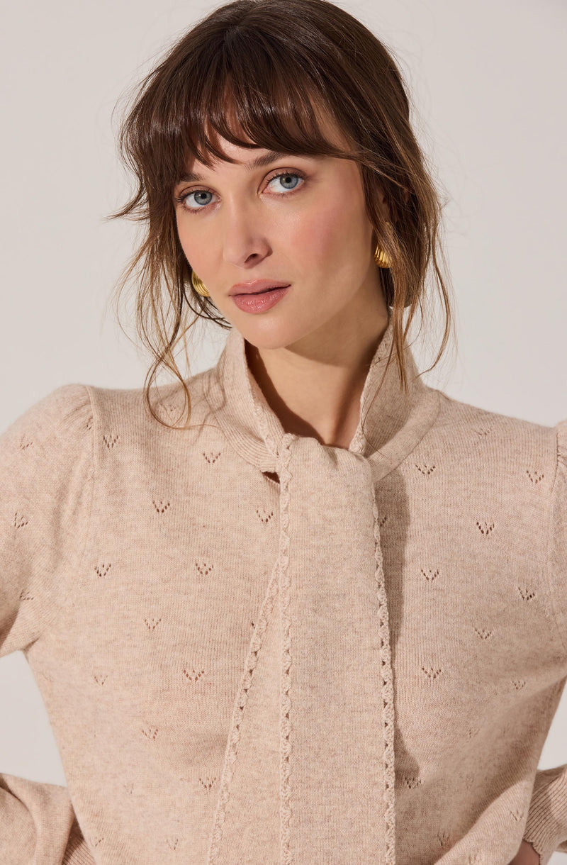 A woman is wearing a beige knit sweater with a subtle pattern of small, V-shaped eyelets throughout. The sweater features a high, banded collar and a decorative vertical trim running down the front.