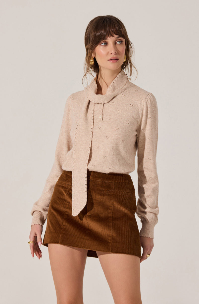 A woman is wearing a cream-colored, long-sleeved sweater with a textured pattern and a matching scarf that drapes down the front. She is also dressed in a high-waisted, brown corduroy mini skirt. She has shoulder-length brown hair with bangs and is accessorized with large gold hoop earrings and rings on her fingers.