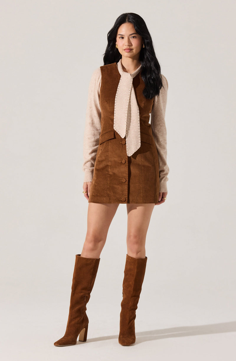 A woman is wearing a beige long-sleeve sweater with a textured, knitted beige tie at the collar. Over the sweater, she has on a sleeveless brown corduroy dress with large front buttons and a fitted waist. She completes her outfit with knee-high brown suede boots with high heels.
