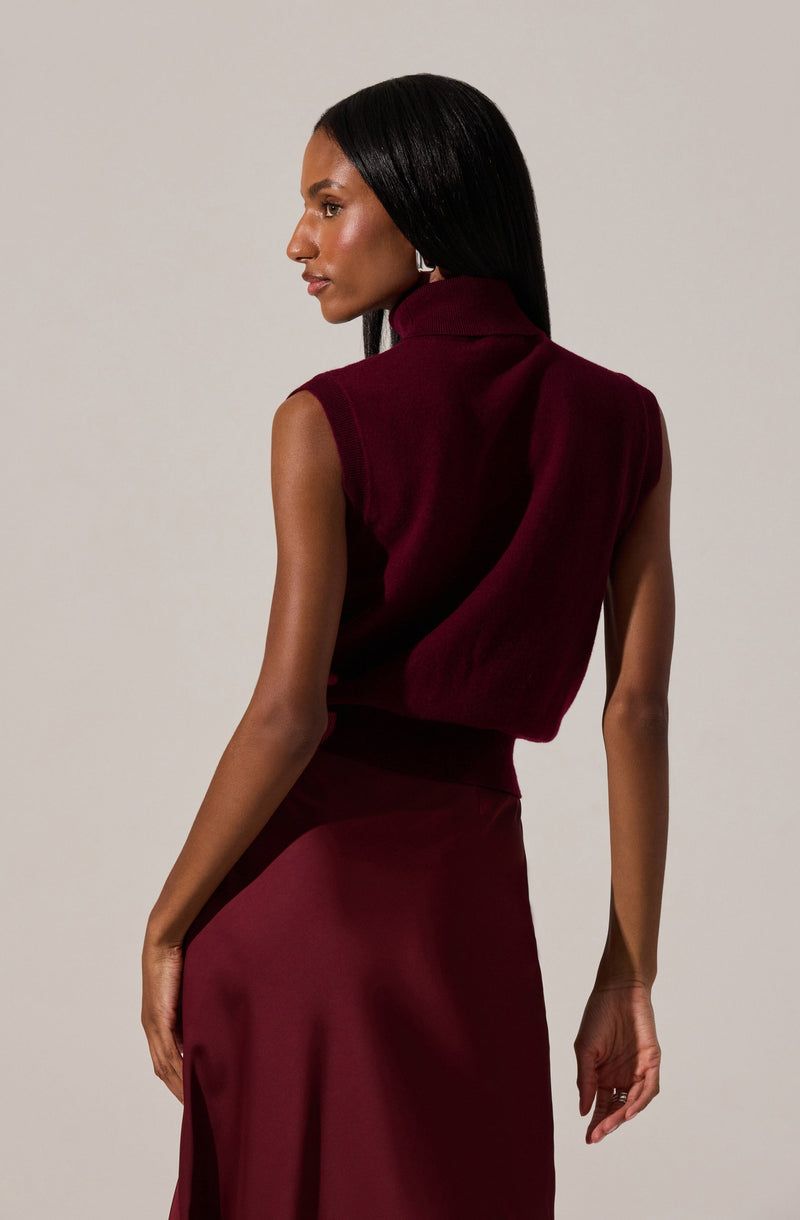 A woman is wearing a sleeveless, high-neck burgundy top with a slightly loose fit. She pairs it with a matching high-waisted skirt that falls below the knee, made of a smooth, flowing fabric.