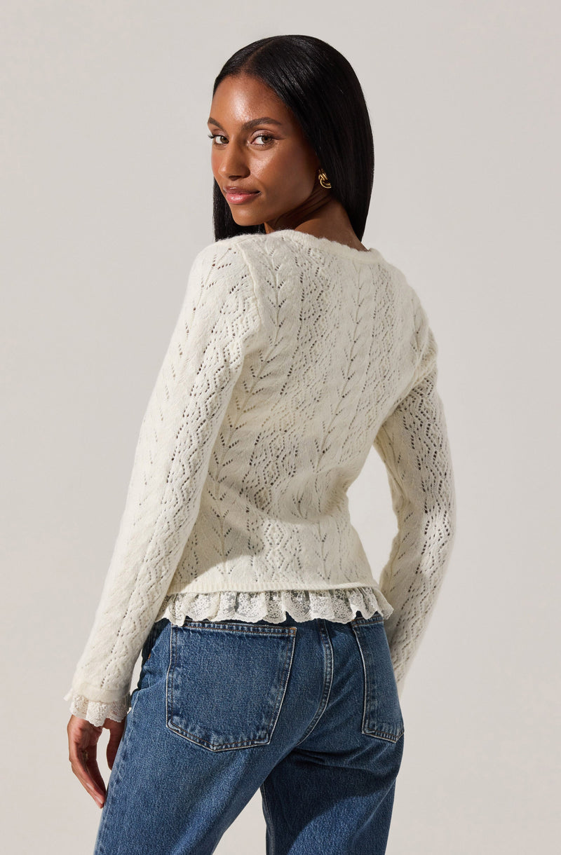 A woman is wearing a cream-colored knit sweater with a textured, open-weave pattern. The sweater has long sleeves, a round neckline, and a ruffled hem at the waist. She pairs it with high-waisted blue jeans.