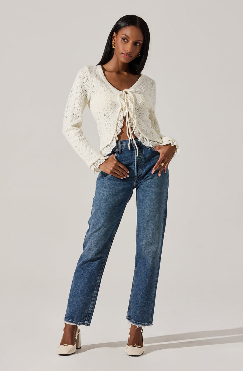 A woman is wearing an off-white, long-sleeved, knitted cardigan with a scalloped hem and ruffled cuffs, featuring a tie-front closure. She pairs it with high-waisted, straight-leg blue jeans and beige high-heeled shoes. She is also wearing gold earrings and a gold ring.