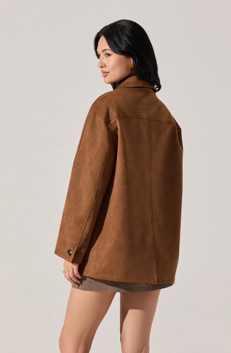 A woman is wearing a loose-fitting, brown suede jacket with a collar and long sleeves. The jacket features buttoned cuffs and appears oversized. She is also wearing a short skirt with a small checkered pattern.