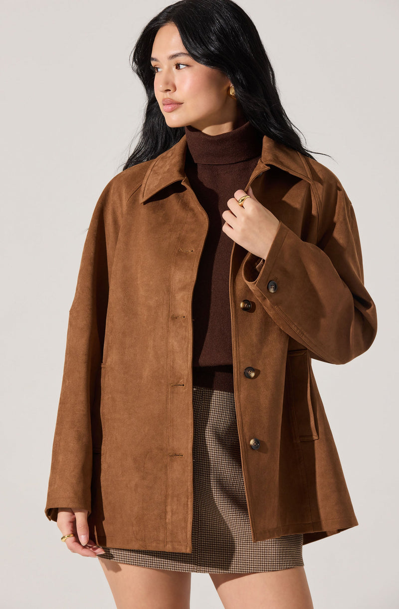 A woman wearing a tan, oversized, button-up jacket with a collar. Underneath, she has on a dark brown turtleneck sweater. She is also dressed in a checkered skirt with a small, subtle pattern.