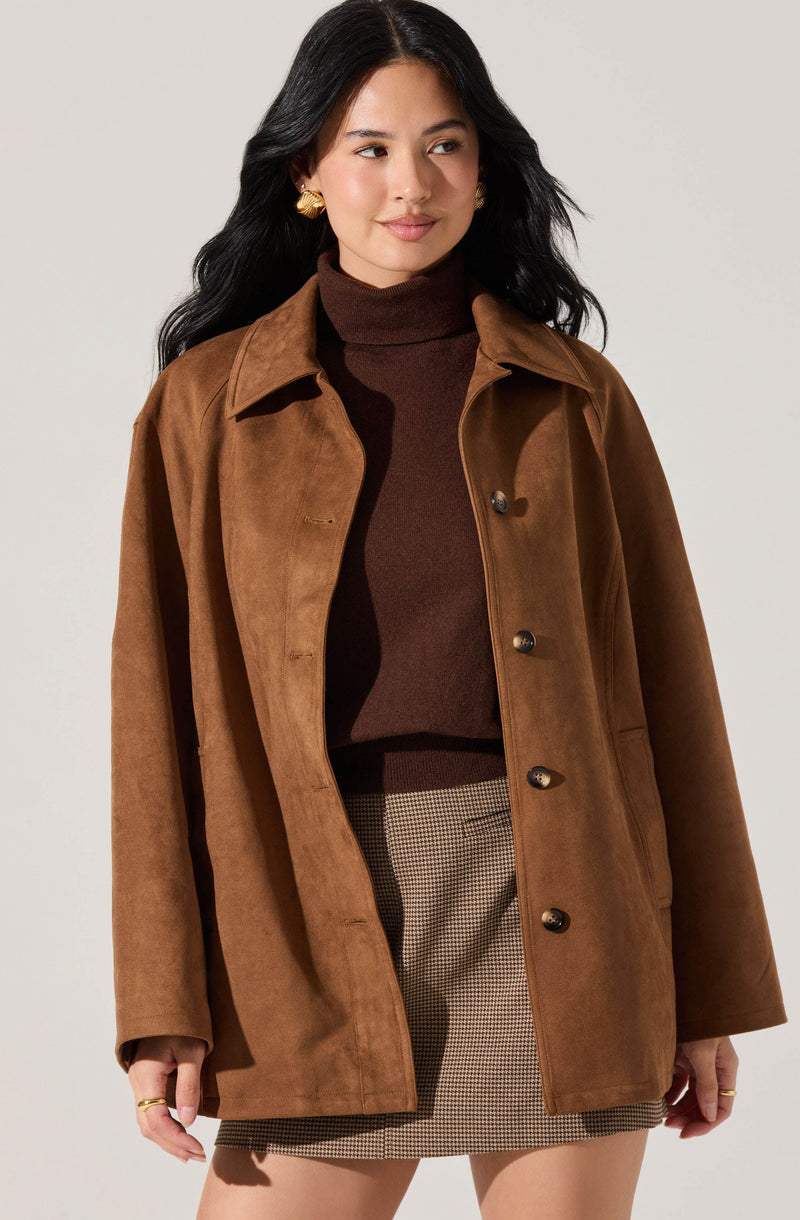 A woman is wearing a brown suede button-up jacket over a dark brown turtleneck sweater. She has paired the outfit with a knee-length checkered skirt. She also has gold earrings and rings on her fingers.