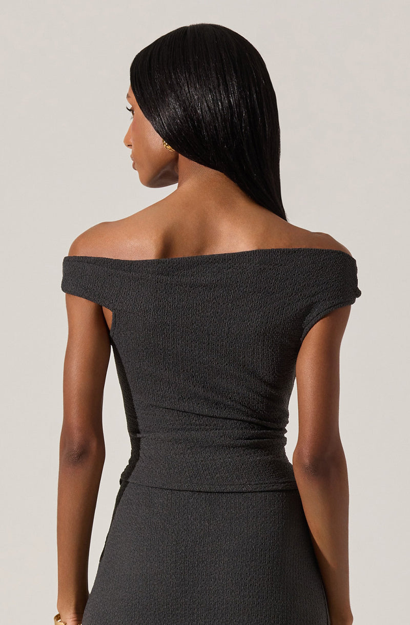 A woman is wearing a form-fitting, off-the-shoulder black dress with a textured fabric. The dress has a wide neckline that drapes over her shoulders, exposing her collarbones and upper back.