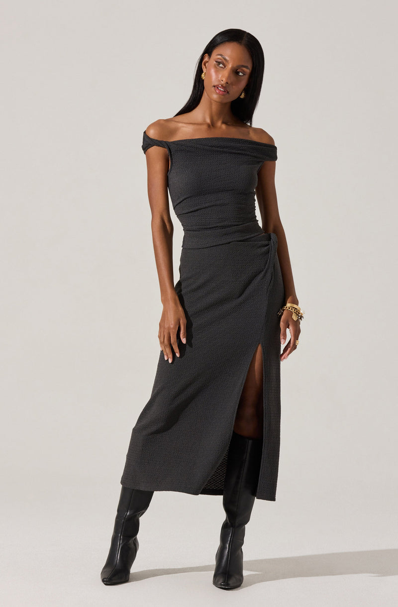A woman is wearing an off-the-shoulder black dress with a textured fabric. The dress has a slit up the front, revealing one leg, and drapes slightly at the waist. She pairs the dress with tall black boots and accessorizes with gold jewelry, including earrings and bracelets.