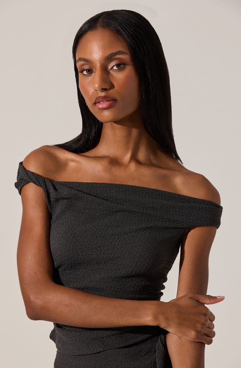 A woman wearing an off-the-shoulder black top with a textured fabric. The top has a wide neckline that drapes slightly off her shoulders and short sleeves gathered at the shoulders.