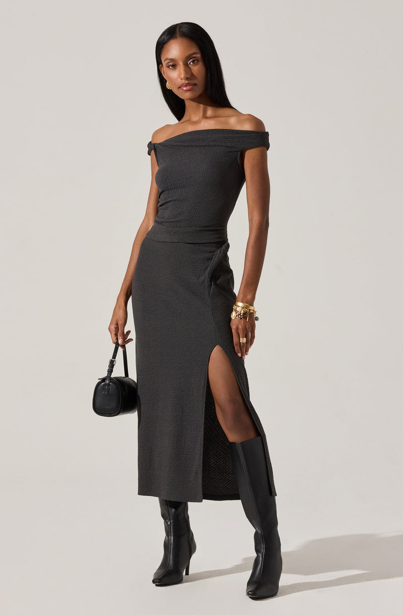A woman is wearing an off-the-shoulder, short-sleeved, fitted black dress with a high slit on the side. She is accessorized with gold jewelry, including earrings and multiple bracelets. She is carrying a small black handbag and wearing knee-high black boots.