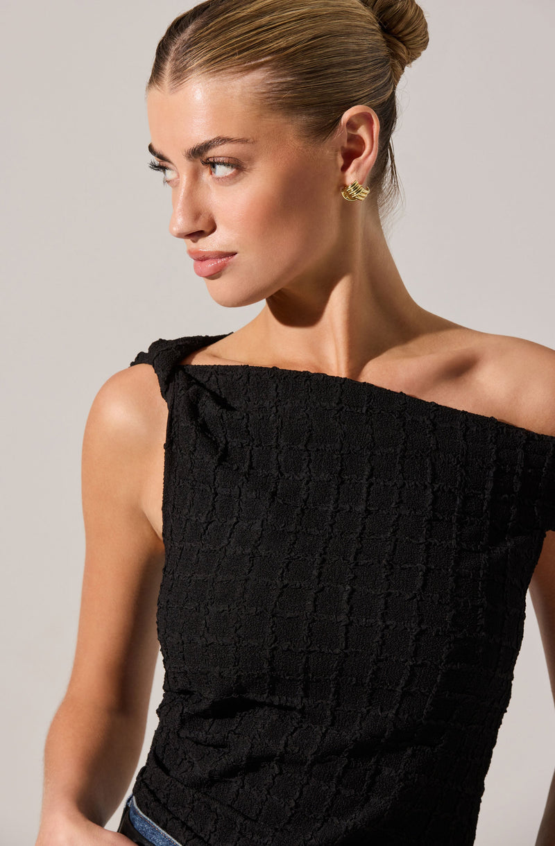A woman is wearing a sleeveless black textured top with a wide, off-the-shoulder neckline. She has gold earrings, her hair is styled in a sleek bun, and she has a neutral makeup look.