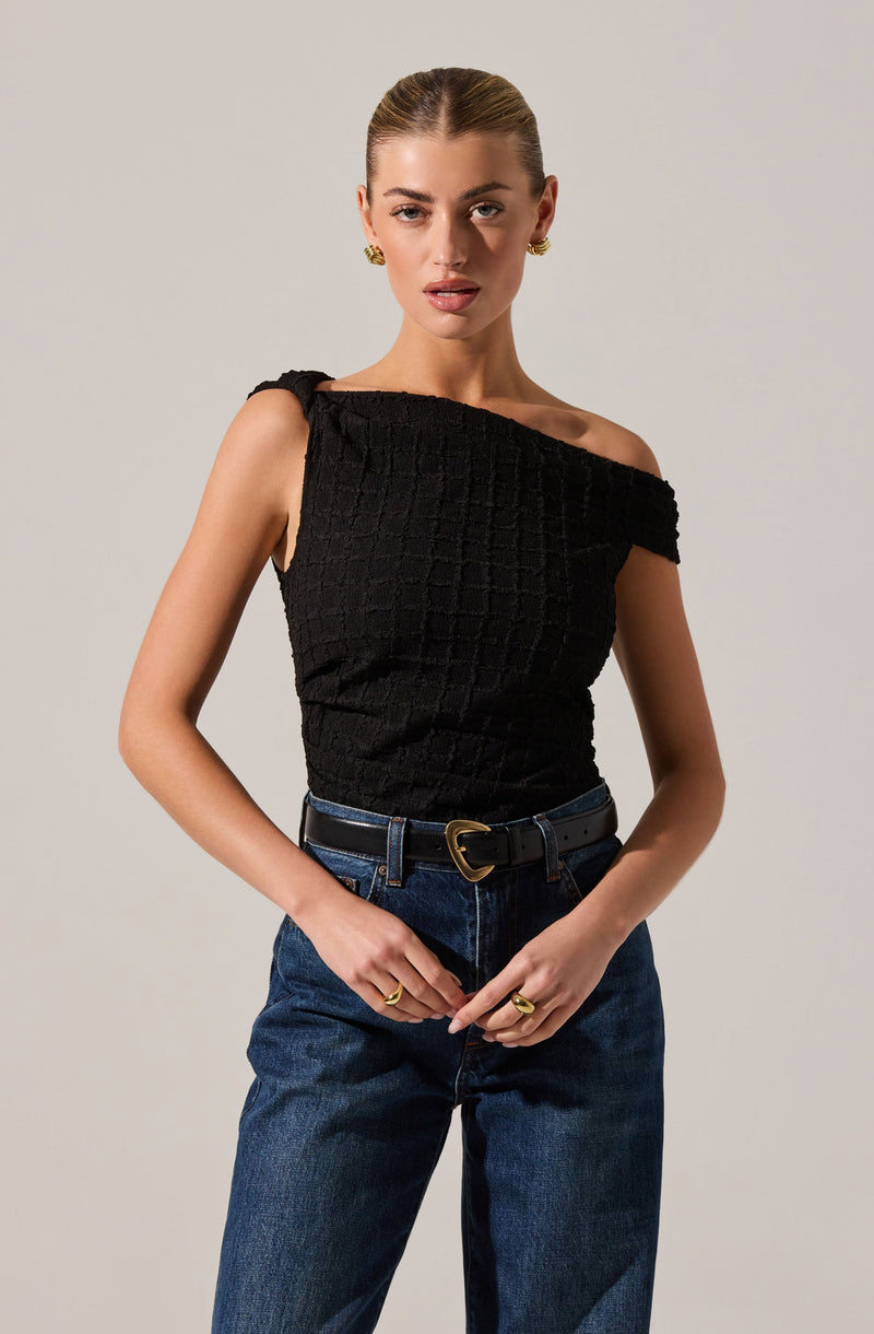 A woman is wearing a black sleeveless top with a textured, grid-like pattern and an asymmetrical, off-shoulder neckline. She pairs the top with high-waisted blue jeans and a black belt featuring a gold heart-shaped buckle. She accessorizes with gold earrings and multiple rings.