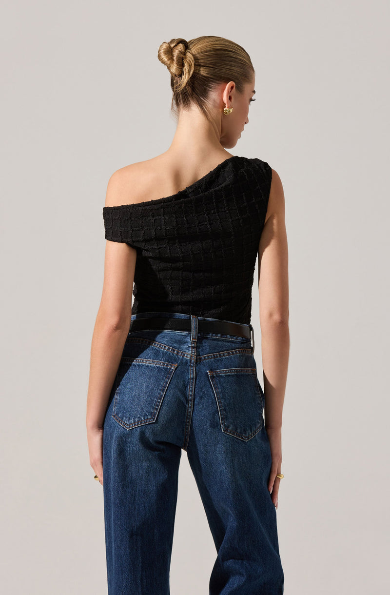 A woman is wearing a black, textured, off-the-shoulder top paired with high-waisted blue jeans. The top has a square-patterned appearance, and the jeans have classic back pockets with orange stitching. She accessorizes with gold earrings and a ring. Her hair is styled in a sleek, low bun.
