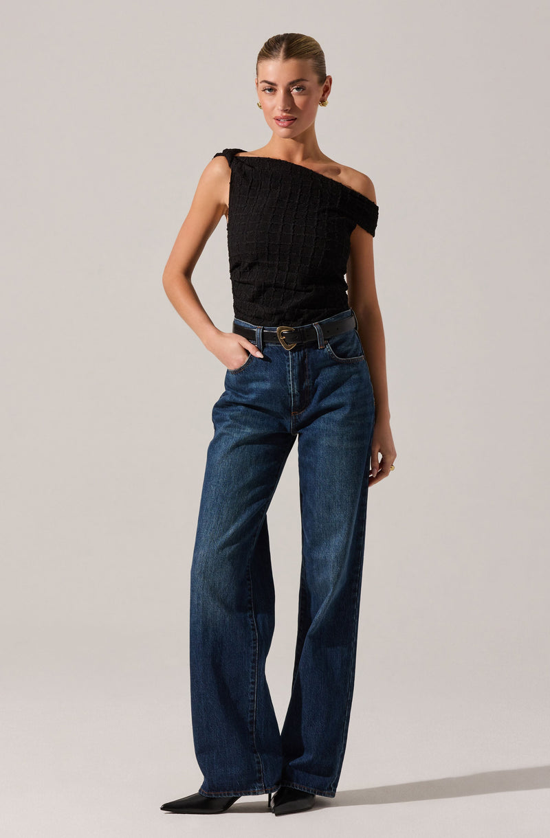 A young woman is wearing a black, sleeveless, asymmetrical top with textured fabric that has a one-shoulder design. She pairs it with high-waisted, loose-fitting blue jeans and a black belt with a gold buckle. She completes her outfit with pointed black shoes.