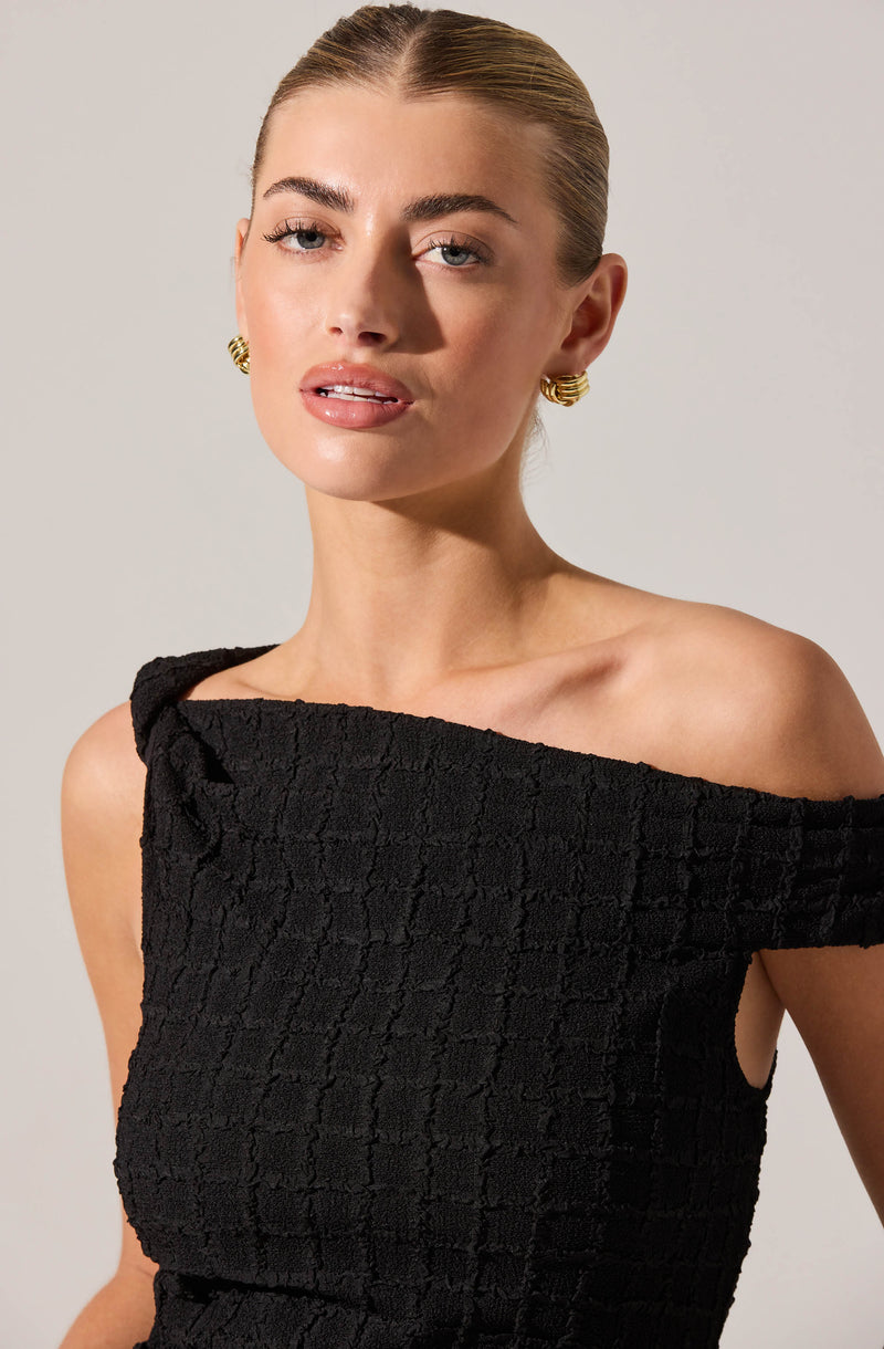 A woman with blonde hair styled in a sleek, pulled-back look is wearing a black, textured, off-the-shoulder dress. The dress has a square neckline and a unique shoulder strap on one side, revealing her collarbone and shoulder. She accessorizes with gold spiral earrings. The background is plain and light-colored.
