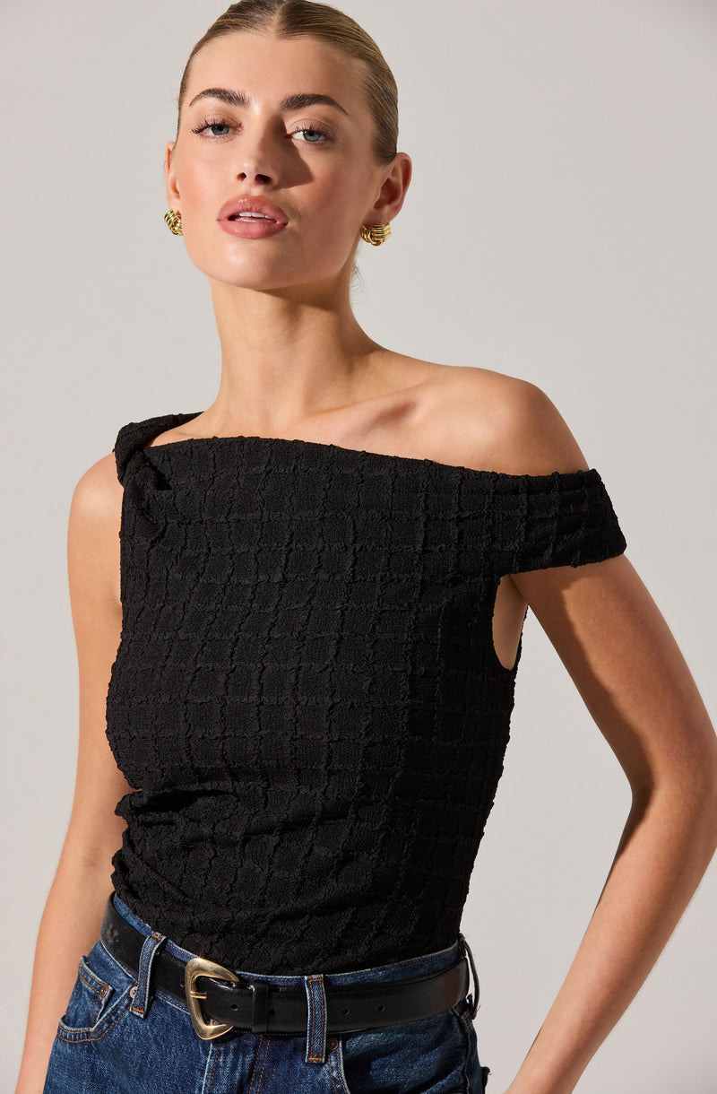 A woman is wearing a black off-the-shoulder top with a textured, checkered pattern. The top has a sleeve that drapes over one shoulder, leaving the other shoulder bare. She pairs the top with blue jeans and a black belt with a gold buckle. She has gold earrings and her hair is slicked back.