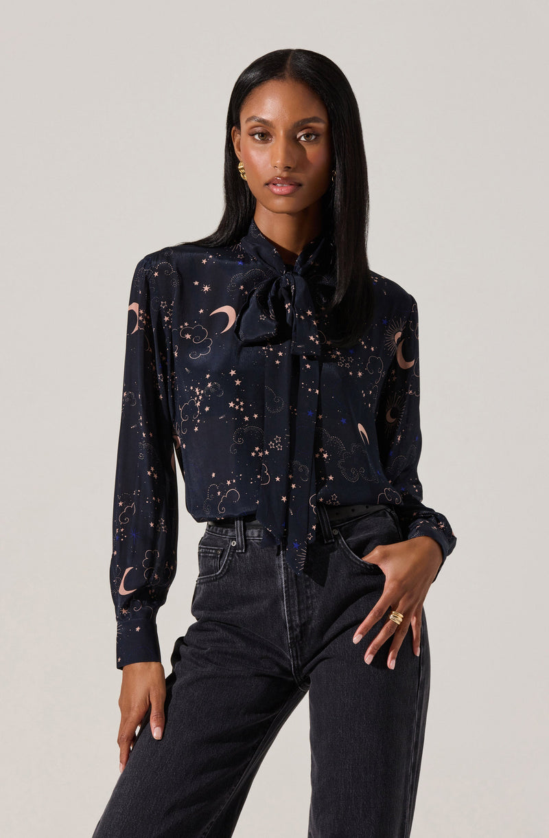 A woman is wearing a long-sleeved, dark-colored blouse with a celestial pattern that includes moons, stars, and clouds. The blouse features a bow tie at the collar. She has paired it with dark high-waisted jeans.