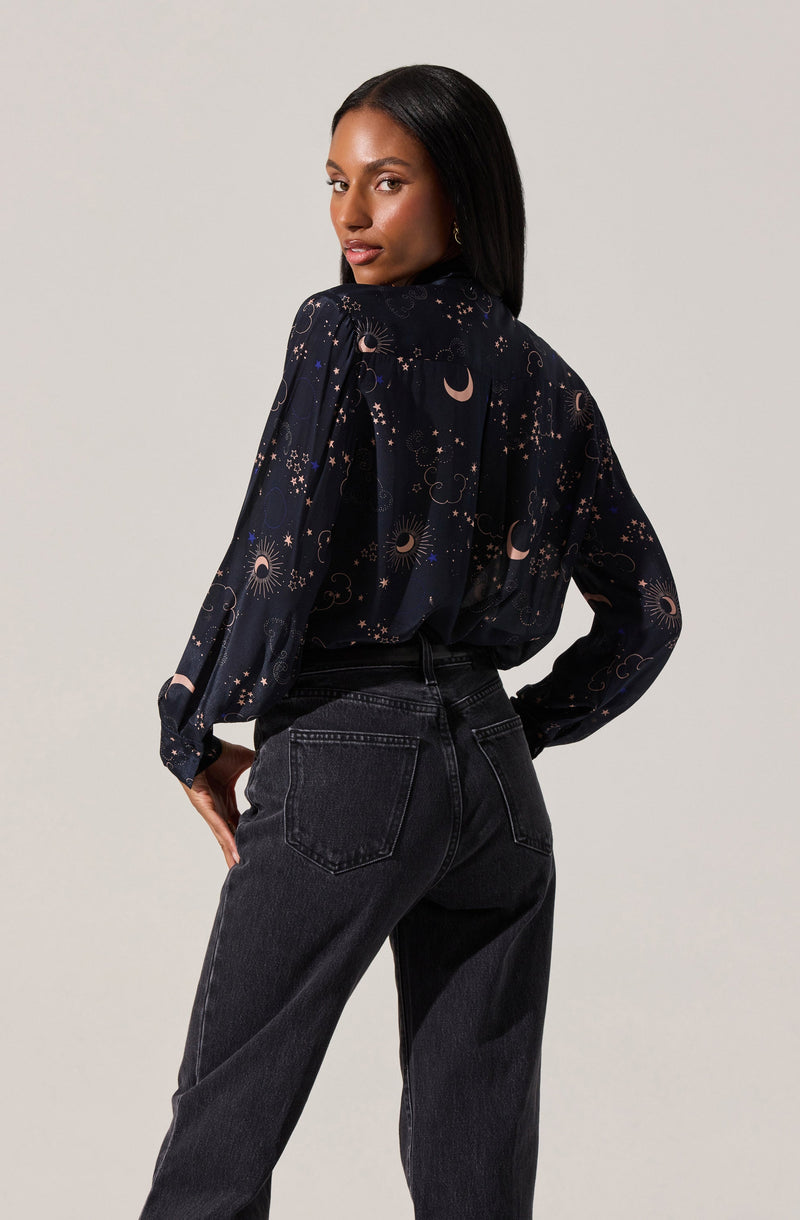 A woman is wearing a long-sleeved blouse with a dark background featuring a celestial print that includes moons, stars, and suns. She pairs it with high-waisted black jeans.