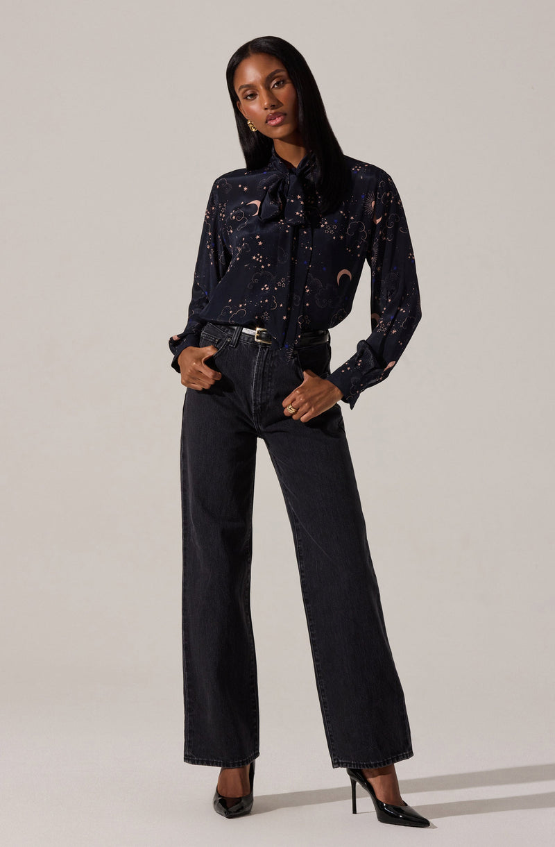 A woman is wearing a dark, long-sleeved blouse with a celestial pattern featuring moons and stars, and a large bow tied at the collar. She pairs the blouse with high-waisted, wide-leg black jeans and black pointed-toe high heels.