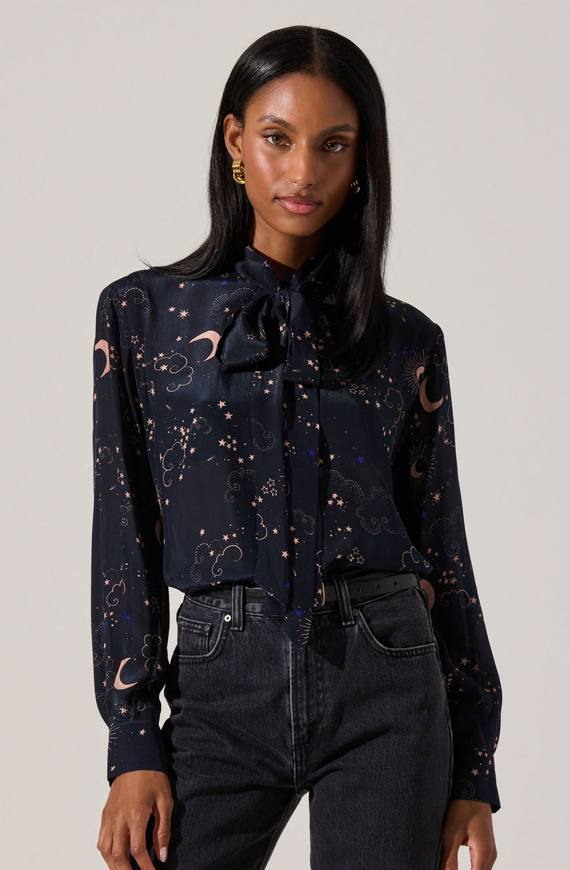 A woman is wearing a black long-sleeve blouse with a celestial pattern featuring moons, stars, and clouds in soft pink and blue hues. The blouse has a high neckline with a large bow tied at the collar. She pairs it with high-waisted black denim jeans.