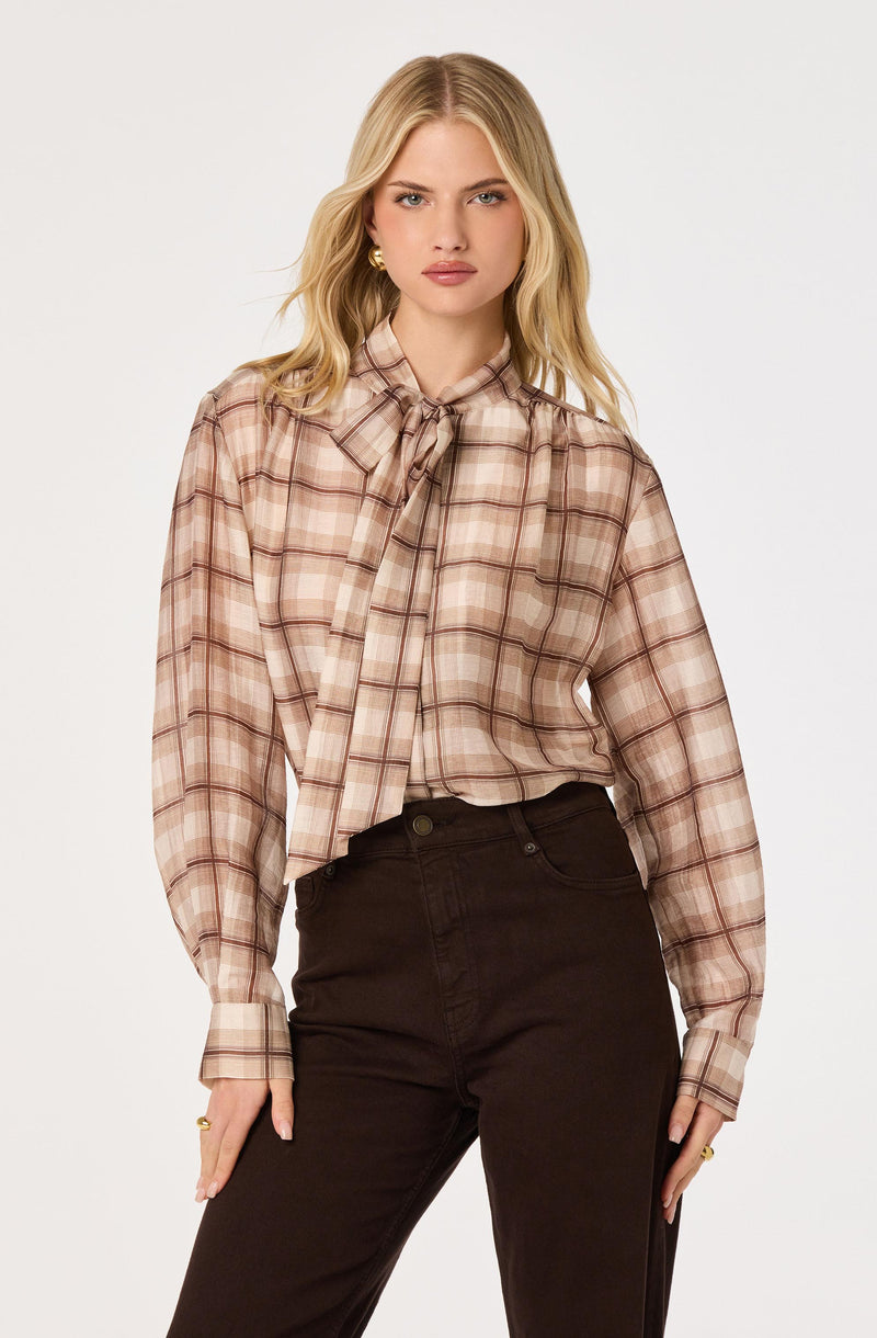 Woman wearing a beige and brown plaid blouse with a bow tie at the collar and long sleeves.