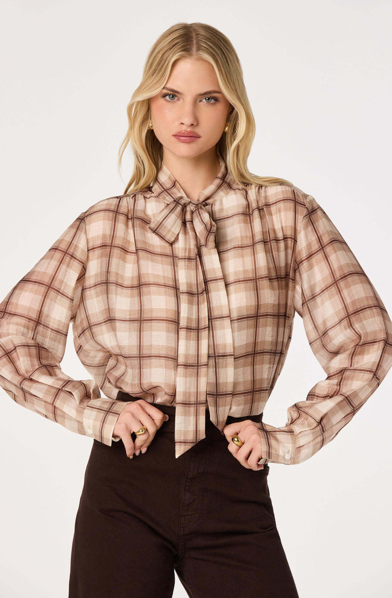 Woman wearing a beige plaid blouse with a bow tie collar and long sleeves.