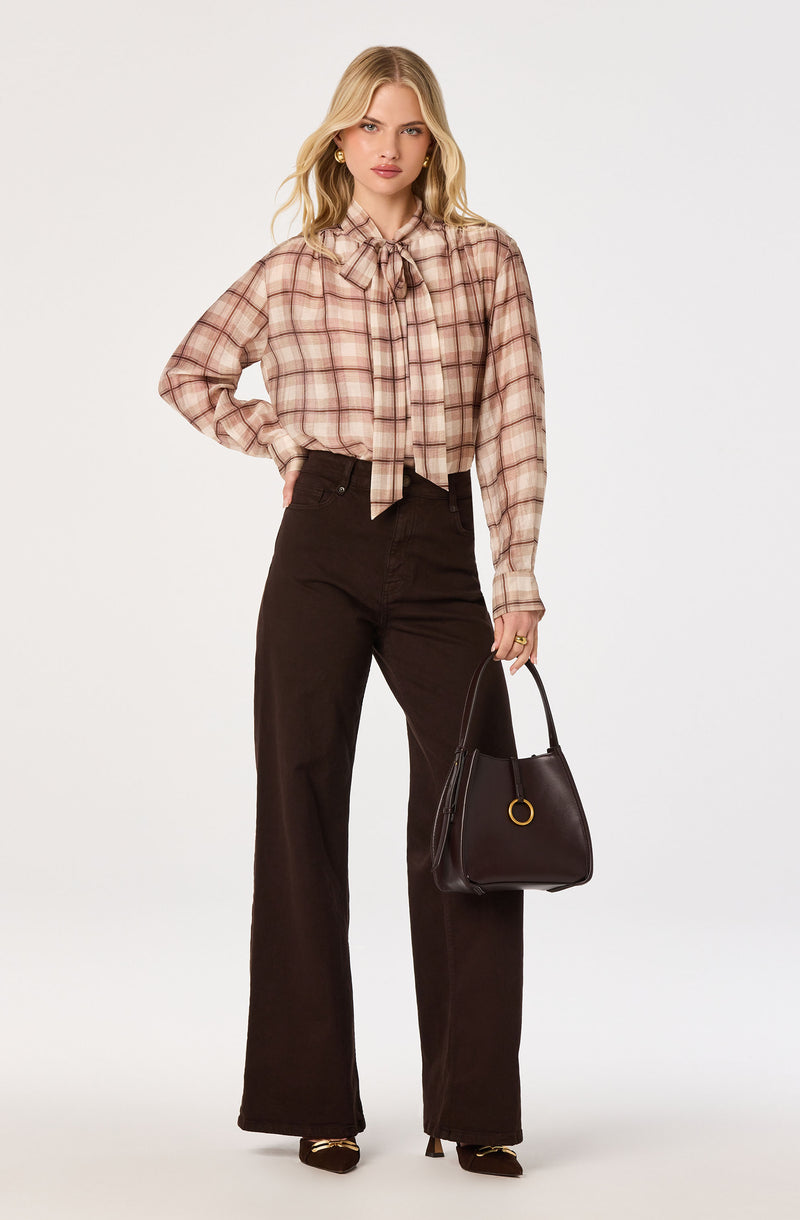 Woman in a plaid beige and pink button-up shirt with a bow tie, paired with black high-waisted wide-leg pants, holding a black handbag.