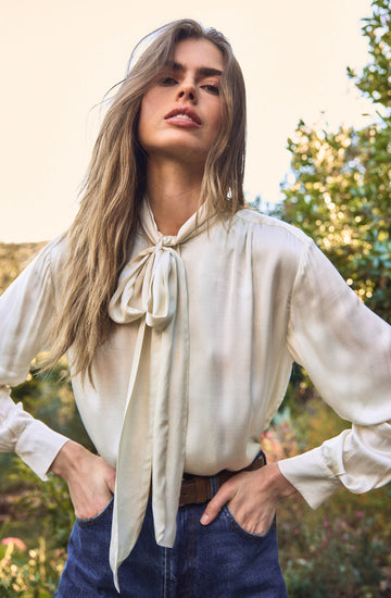 A woman is wearing a cream-colored, satin blouse with long sleeves and a large bow tied at the collar. The blouse has a loose fit with pleats at the shoulders. She is also wearing high-waisted blue jeans with a brown belt.