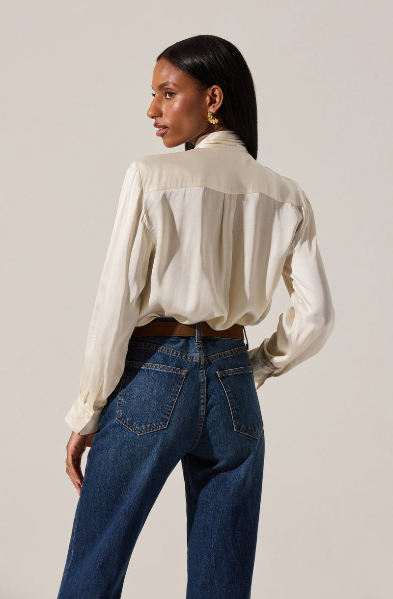 A woman is wearing a long-sleeved, cream-colored blouse with a high collar and pleated details on the back. She pairs it with high-waisted, dark blue denim jeans that have visible stitching and back pockets.