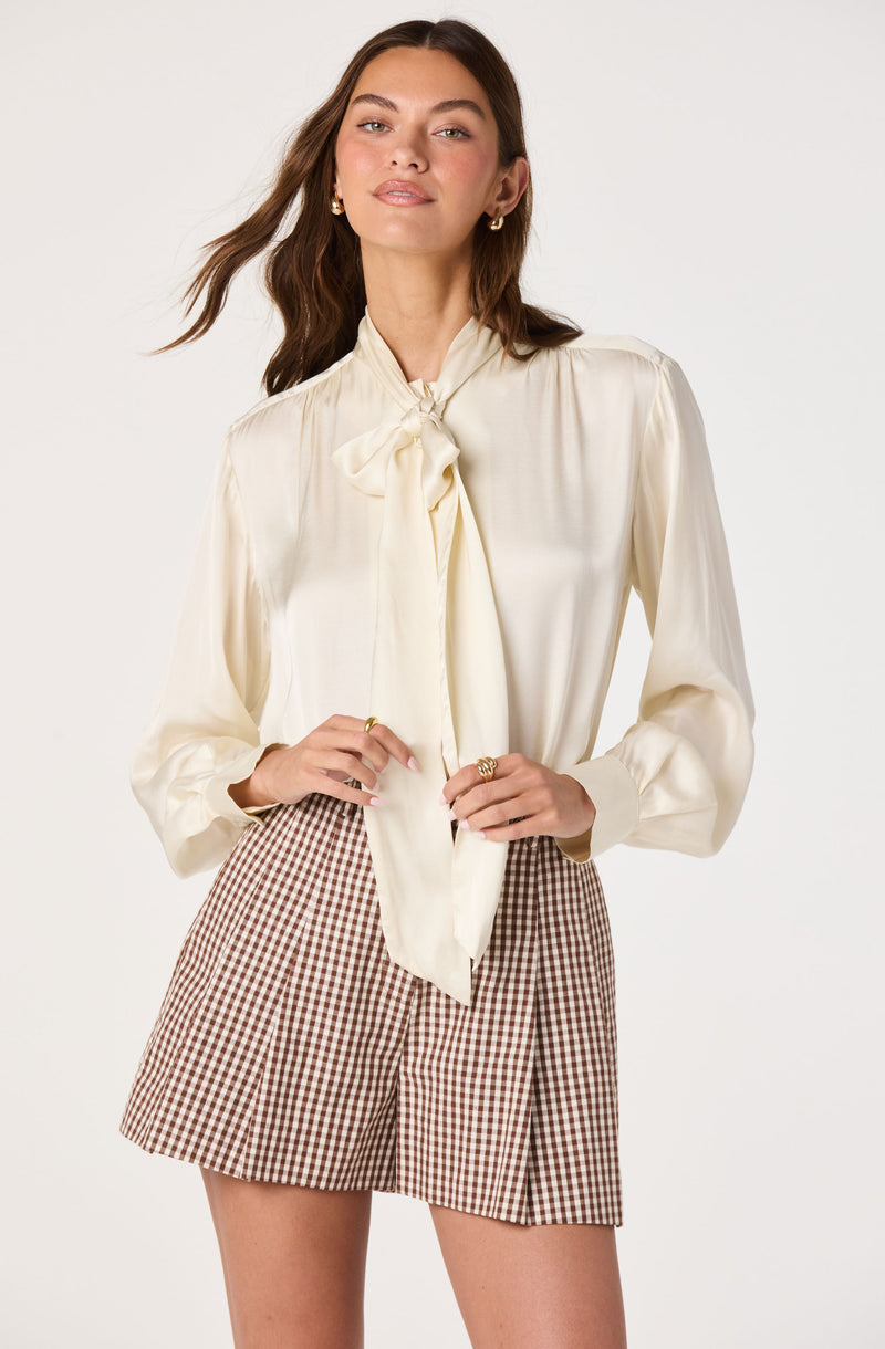 Woman in cream satin blouse with bow tie and checkered shorts.