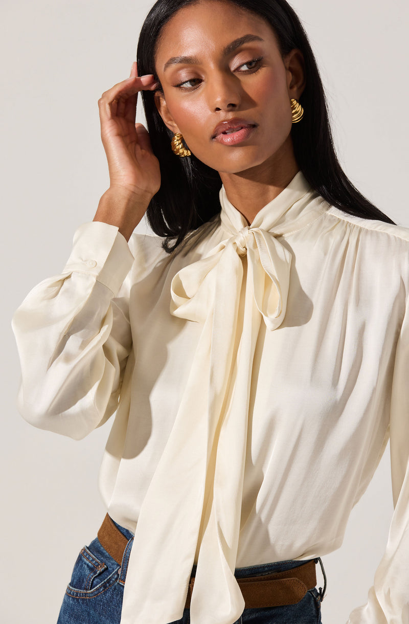 A woman is wearing a cream-colored satin blouse with a large bow tied at the neck. The blouse has long sleeves with buttoned cuffs. She is also wearing gold hoop earrings and blue jeans with a brown belt.