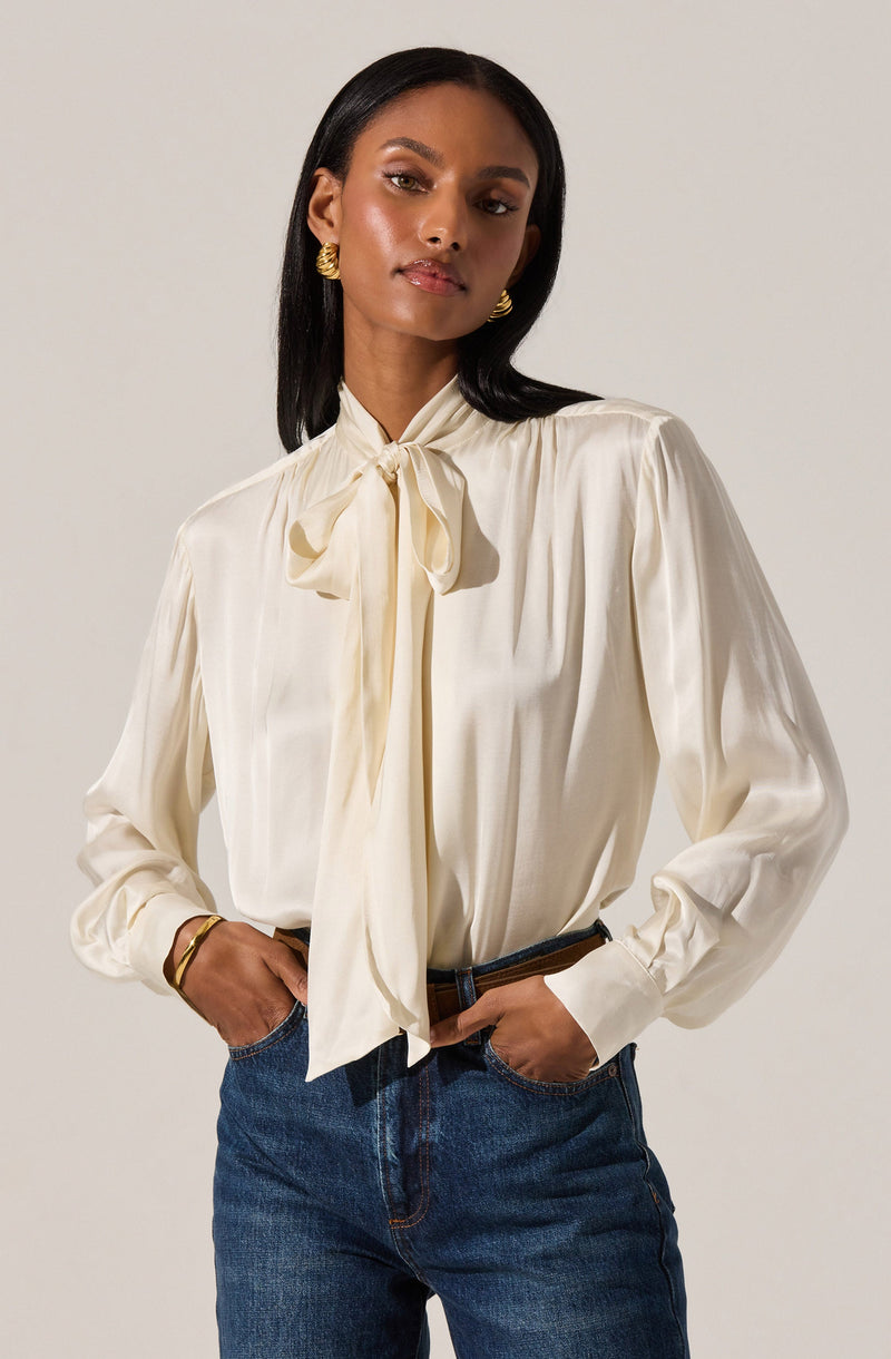 A woman is wearing a long-sleeved, cream-colored blouse with a large bow tied at the neck. The blouse has a silky texture and subtle pleats running down the front. She has paired it with blue jeans and accessorized with gold jewelry, including earrings and a bracelet.