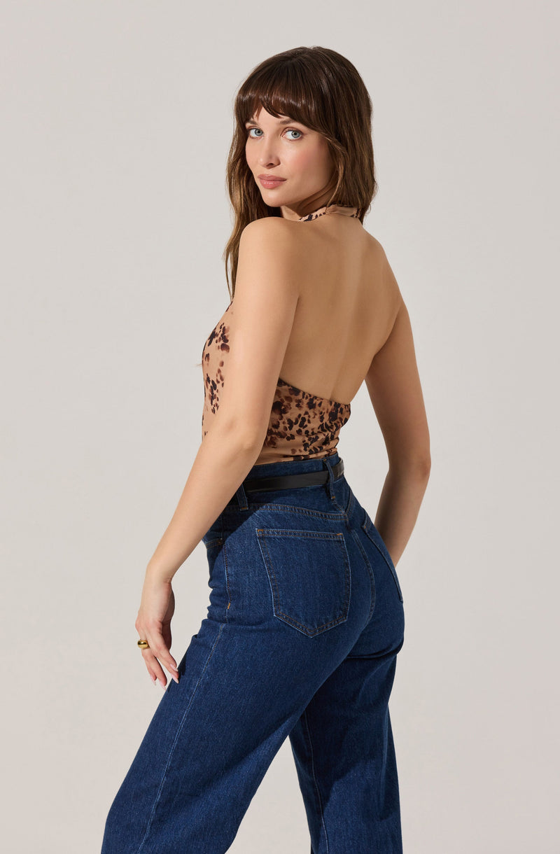 A woman with shoulder-length brown hair and bangs is wearing a halter top with a brown and black leopard print pattern. She is also wearing high-waisted, dark blue denim jeans. She is standing against a plain light gray background, turned slightly to the side with her head turned toward the camera.