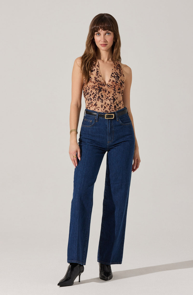 A woman is wearing a sleeveless, V-neck top with a tan background and a black and dark brown leopard print pattern. She pairs it with high-waisted, dark blue straight-leg jeans that have front pockets and belt loops, accessorized with a black belt featuring a gold rectangular buckle. She completes her outfit with black ankle-high pointed-toe stiletto boots.
