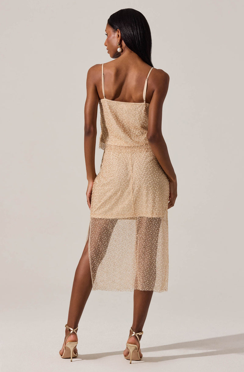 A woman is wearing a two-piece dress with a fitted, glittering gold top with thin spaghetti straps and a matching gold skirt. The skirt is knee-length with an overlay of sheer, glittery fabric that extends below the solid layer, creating a semi-transparent look. The woman is also wearing high-heeled sandals and large pearl earrings.