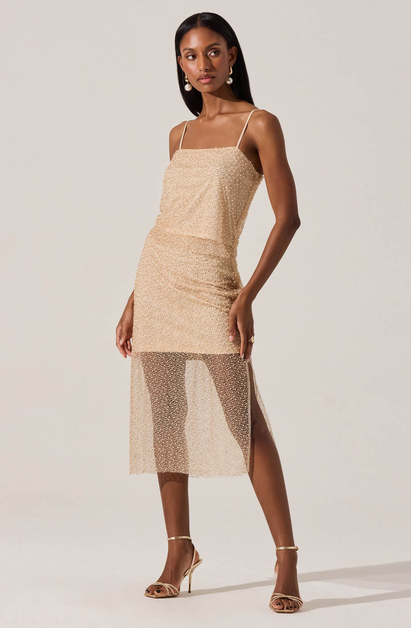 A woman is wearing a beige dress with thin spaghetti straps. The dress has a fitted silhouette with a layered design, featuring a solid upper layer and a sheer, sparkly lower layer that extends to mid-calf length and has a side slit. She is also wearing open-toed, high-heeled sandals in a matching beige tone.