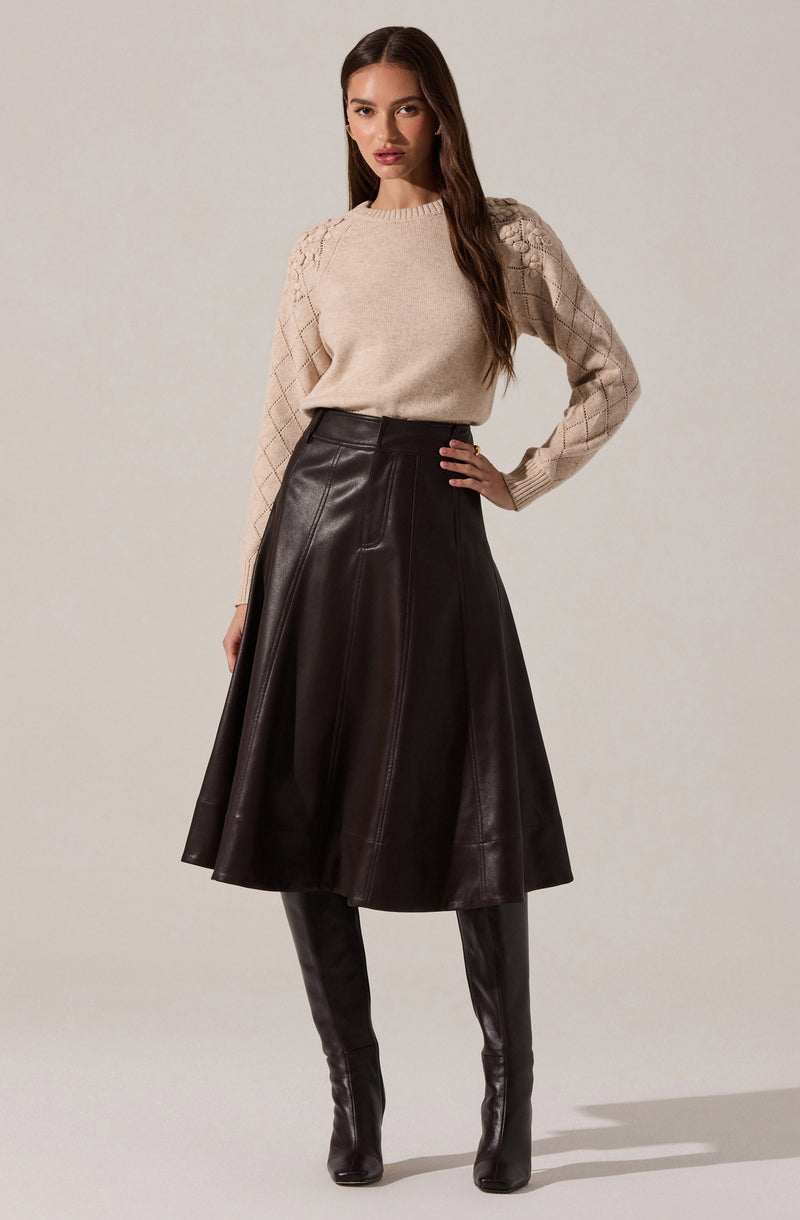 A woman is wearing a beige knit sweater with textured diamond patterns on the shoulders and sleeves. She pairs it with a high-waisted, dark brown leather A-line skirt that falls below the knees. She completes the outfit with tall, black leather thigh-high boots.