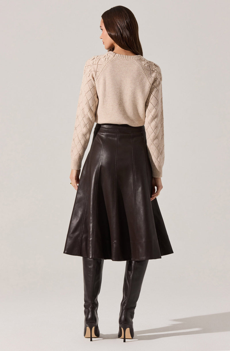 A woman is wearing a light beige sweater with textured diamond patterns on the sleeves and shoulders. She pairs it with a high-waisted, dark brown leather skirt that flares out below the waist. She completes the outfit with tall, matching dark brown leather thigh-high boots with high heels.