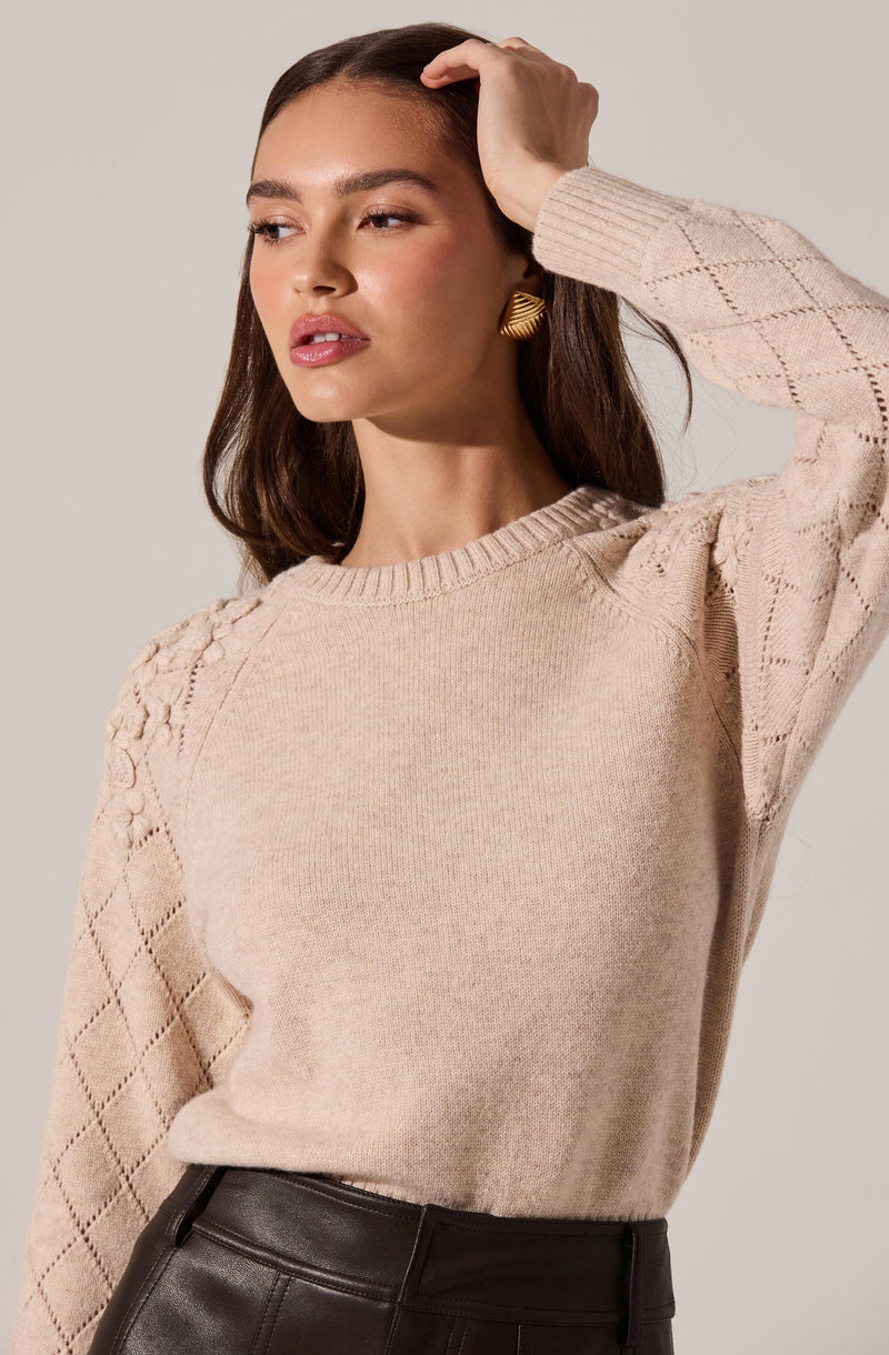 A woman is wearing a beige knitted sweater with diamond-shaped lace patterns on the sleeves and shoulders. The sweater has a round neckline, and she pairs it with high-waisted, dark brown leather pants. She has long, dark hair and is accessorized with a gold spiral-shaped earring.