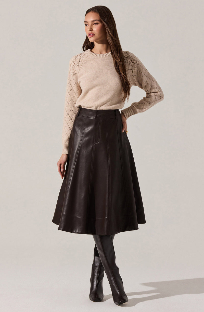 A woman is wearing a beige knit sweater with a textured pattern on the shoulders and sleeves, paired with a high-waisted, knee-length, dark brown leather skirt that has visible stitching details. She completes the look with black knee-high leather boots.